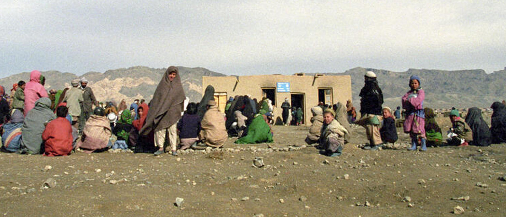 Dozens of refugees, some children, are sitting and standing outside in the dirt in front of a lone small building.